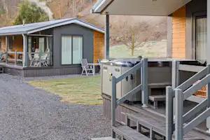 Holz-Ferienhaus mit Terrasse, Liegestühlen und Grill auf dem Vorplatz, im Hintergrund bewaldete Hügel.