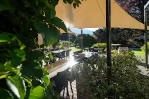 Camping-Terrasse mit Tischen und Stühlen unter einem Sonnenschirm, umgeben von Grün und Blick ins Tal.