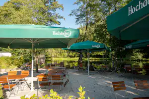 Camping-Terrasse am See mit Holzstühlen und Tischen unter grünen Sonnenschirmen, im Hintergrund Bäume und Wasser.