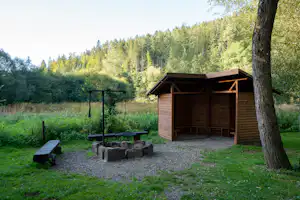Holzhütte am Campingplatz mit Grillrost, Feuerstelle aus Steinen und Bank im grünen Garten, umgeben von Wald.