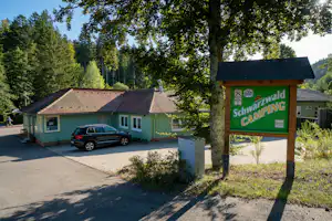 Eingangsschild am Campingplatz Schwarzwald Camping neben einem Parkplatz mit Auto und grünen Holzhäusern im Wald.