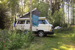 Weißes Wohnmobil steht auf einem Campingplatz im Wald, mit Markise und Vorzelt neben Bäumen und Gras.