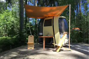 Glamping-Zelt auf Holzterrasse im Wald mit Sonnensegel, Tisch und Stühlen, daneben eine Holzbox und ein Liegestuhl.