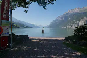 Seeufer mit Badehinweis am Geländer, Kiesstrand und ruhigem Wasser vor grünen Bergen und Felswänden.