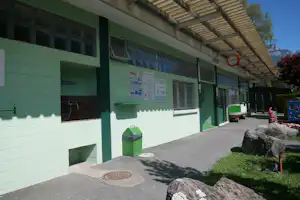 Gebäudewand am Campingplatz mit grünen Türen, Fenstern, Infotafeln und Schildern, daneben Mülltonne und Gehweg.