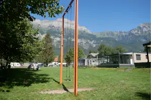 Campingwiese mit Stellplätzen, Wohnwagen und Mobilheimen, im Hintergrund Berge und ein grüner Baum.