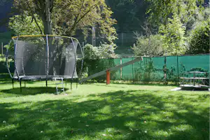 Trampolin im Garten mit grünem Netz, Rasenfläche, Holzrampe und Bäumen im Hintergrund.