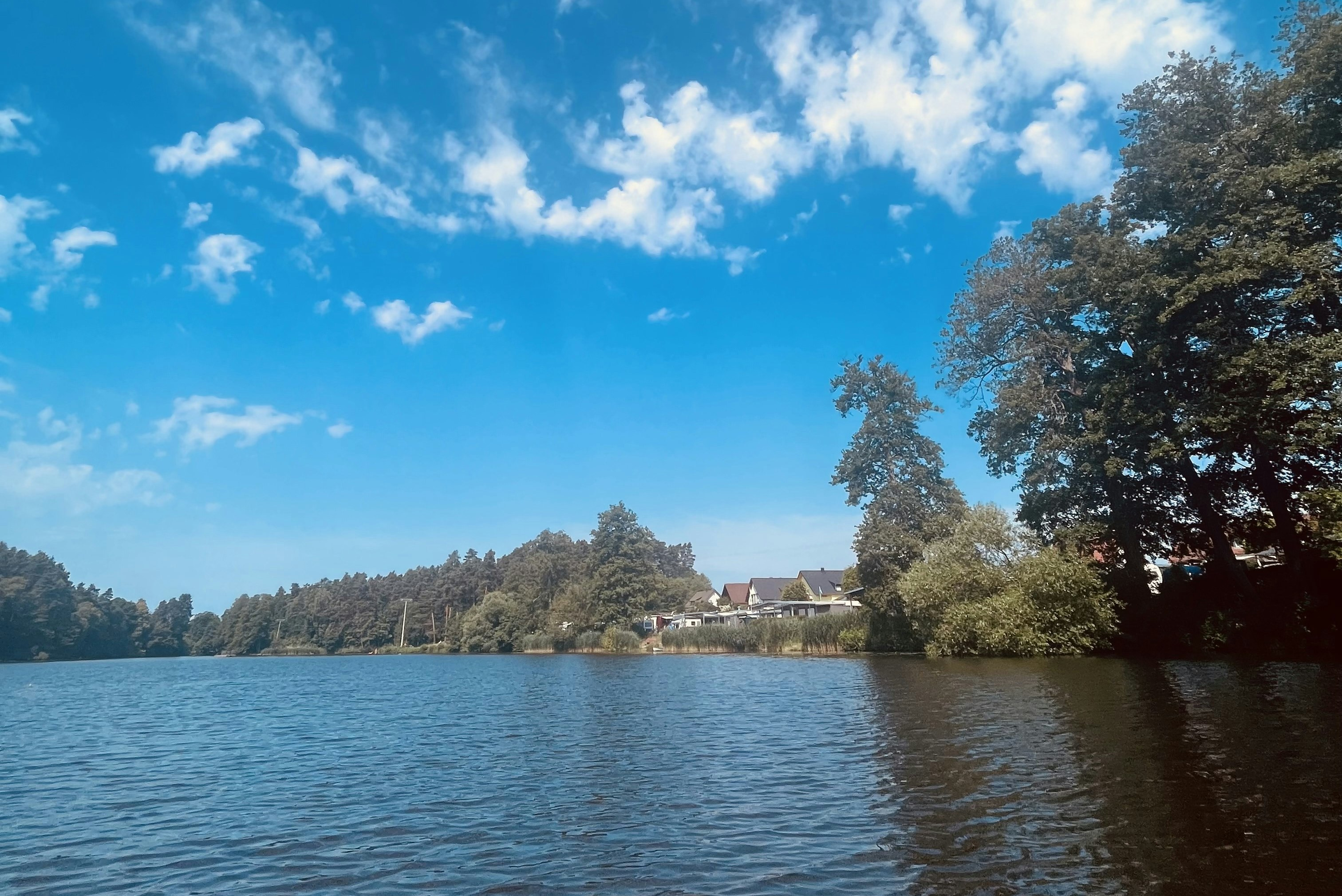 Ruhiger See mit Uferbäumen und Häusern am Horizont, blaues Himmelbild mit Wolken über dem Wasser.
