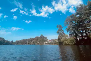 Ruhiger See mit Uferbäumen und Häusern am Horizont, blaues Himmelbild mit Wolken über dem Wasser.