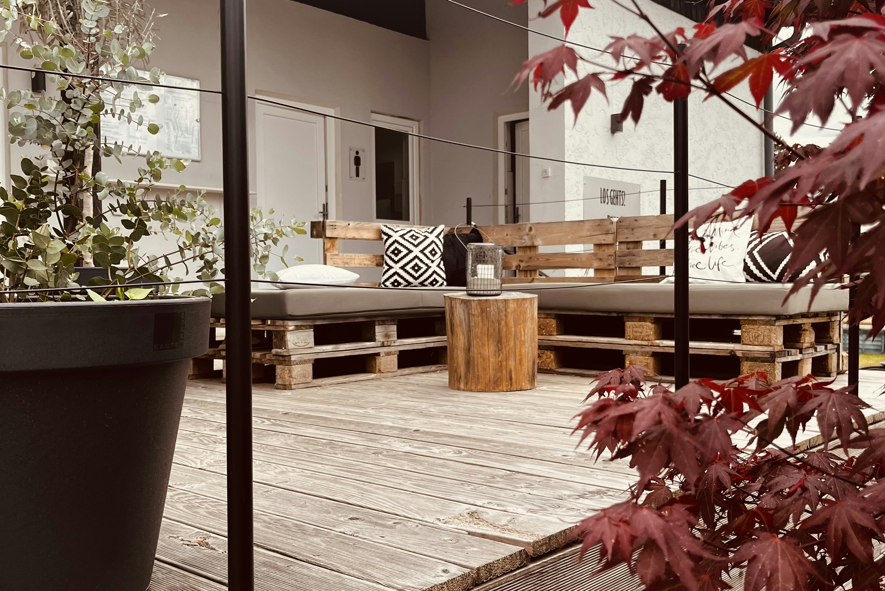 Holzterrasse mit Sitzbank und Pflanzkübeln, im Hintergrund ein Wohnbereich mit Türen und Fensterfront.