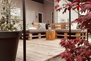 Holzterrasse mit Sitzbank und Pflanzkübeln, im Hintergrund ein Wohnbereich mit Türen und Fensterfront.