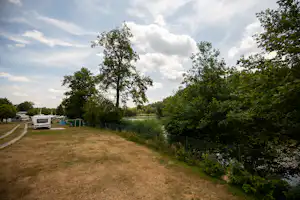 Campingwiese am Fluss mit Bäumen und Wohnwagen am Rand, schmaler Weg entlang des Ufers.