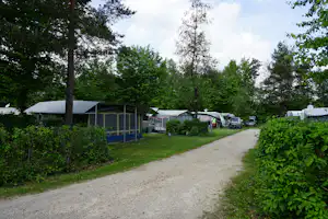 Campingplatzweg mit Wohnwagen und Zelten, Hecken und Bäumen, daneben ein Gebäude mit Markise.