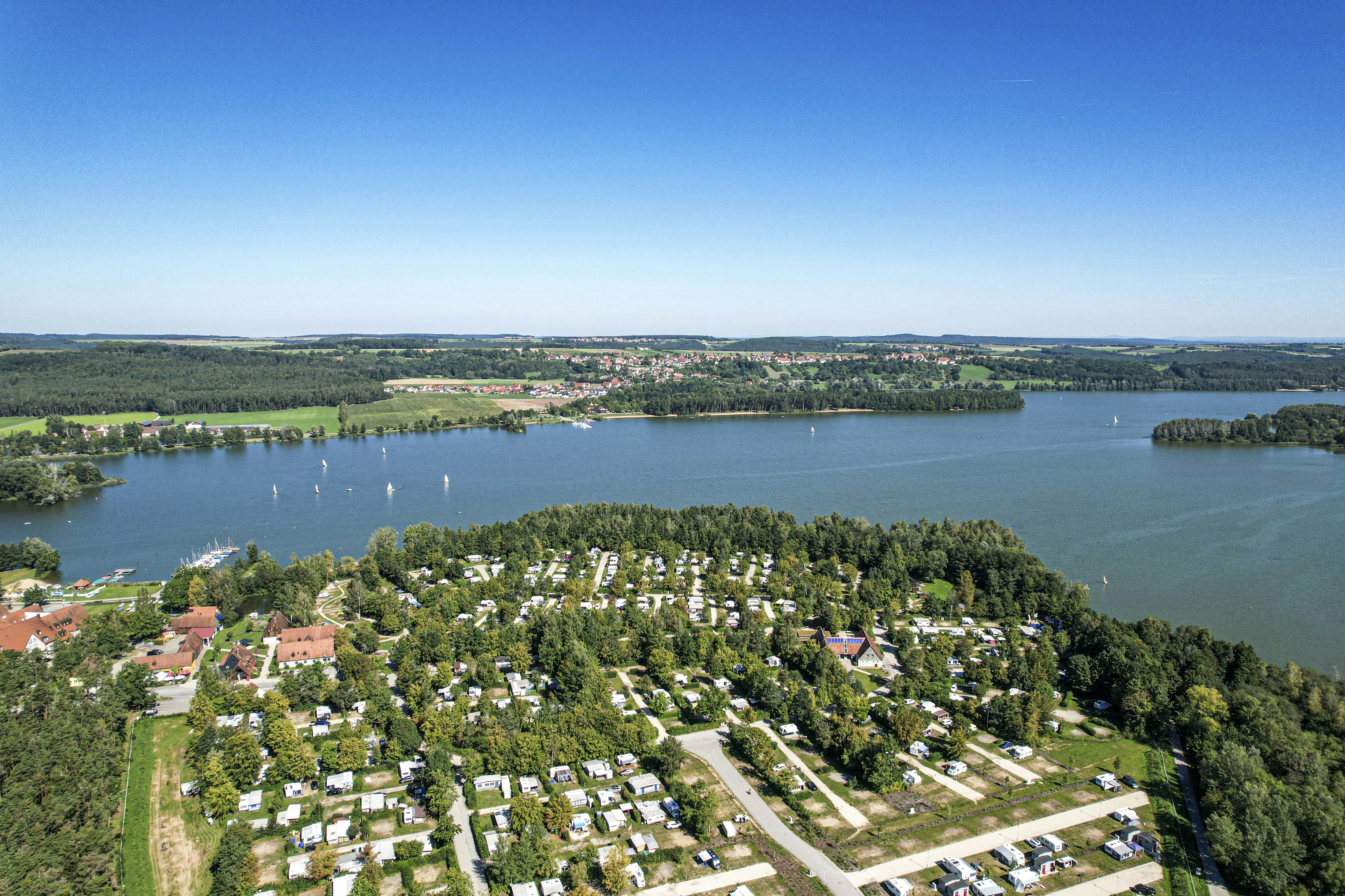 Luftaufnahme eines Campingplatzes am See mit Wohnwagen, Stellplätzen, Wegen und vielen Bäumen am Ufer.
