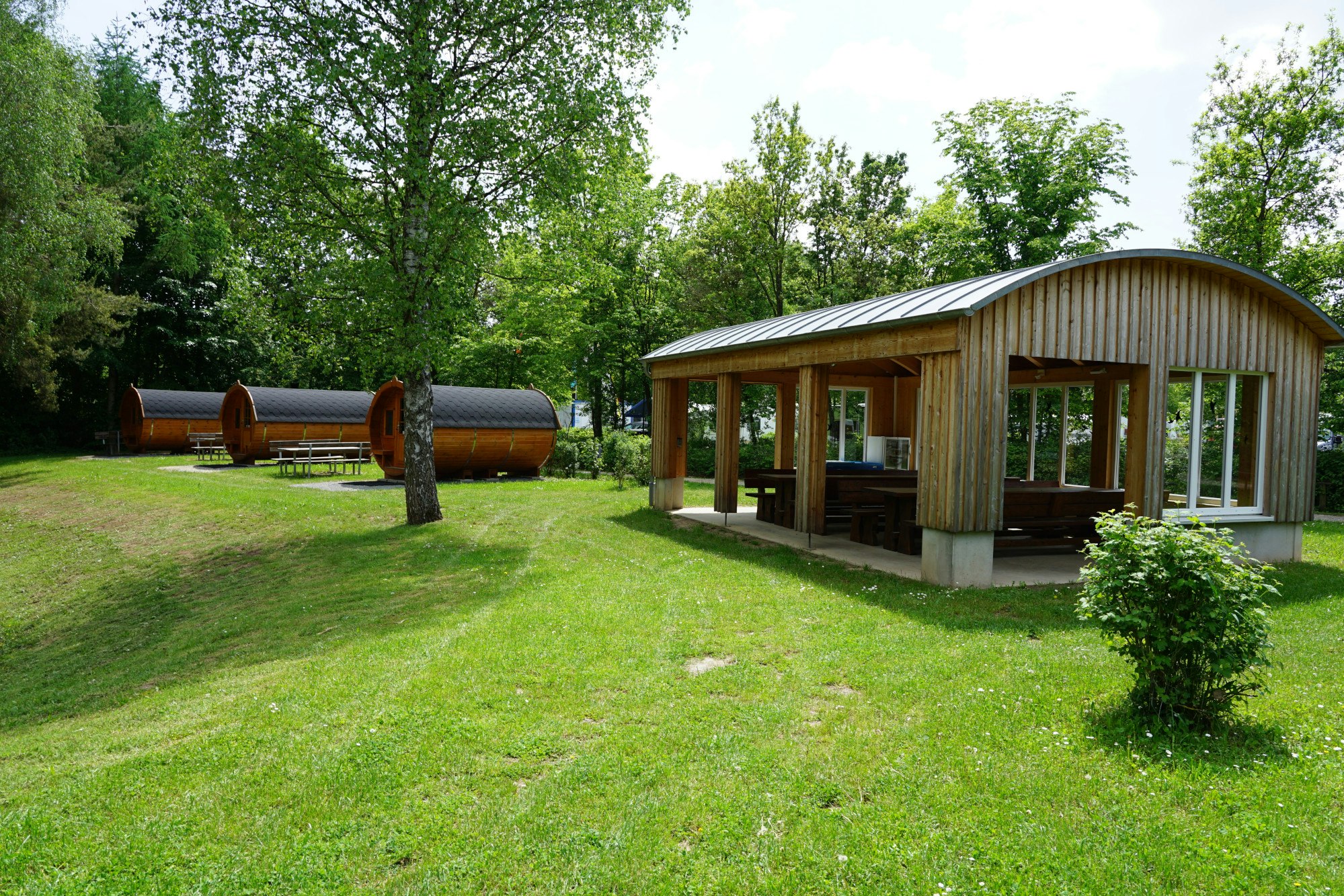 Holzsauna mit Terrasse und Sitzbank auf einer grünen Wiese, daneben ein Holzwohnwagen und Bäume im Hintergrund.