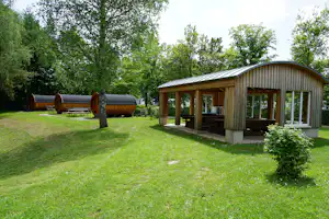 Holzsauna mit Terrasse und Sitzbank auf einer grünen Wiese, daneben ein Holzwohnwagen und Bäume im Hintergrund.