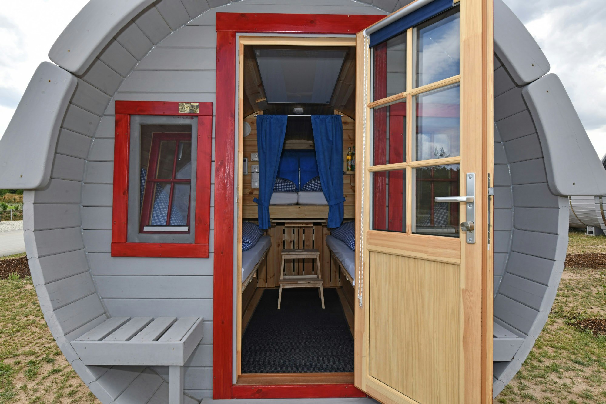 Offener Glamping-Pod-Eingang mit Veranda, Holzbank und großen Fenstern, weißem Rundbau und Rasen im Hintergrund.