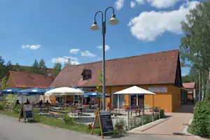 Terrasse am Campingplatz mit Tischen, Stühlen und blauen Sonnenschirmen vor Gebäuden mit roten Ziegeldächern.