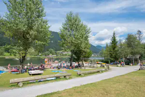Badebereich am See mit Liegen, Handtüchern und vielen Menschen im Wasser, umgeben von Bäumen und Bergen.