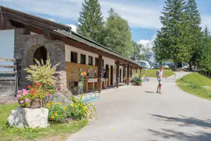 Außenküche am Campingplatz mit Steinwand, Holzüberdachung und Blumenbeeten am Weg, im Hintergrund Wohnmobile.