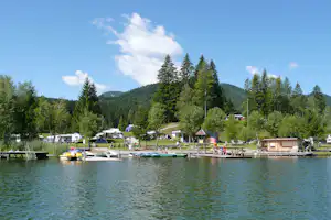 Campingplatz am See mit Booten am Steg, Wohnmobilen und Zelten im Grünen, Bäume und Berge im Hintergrund.