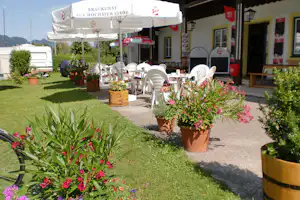 Terrasse am Campingplatz mit weißen Stühlen, Sonnenschirmen und Blumen in Töpfen vor dem Gebäude.