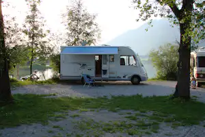 Wohnmobil am Seeufer im Grünen, mit Markise, Campingstuhl und Fahrrad am Stellplatz zwischen Bäumen.