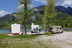 Wohnmobile am See auf einem Stellplatz, mit Fahrrädern, Tischen und Stühlen vor dem Bergpanorama.