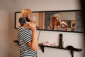 Frau mit blondem Haar spricht in ein Mikrofon vor einem Spiegel im Innenraum, daneben ein Regal mit Flaschen und Deko.