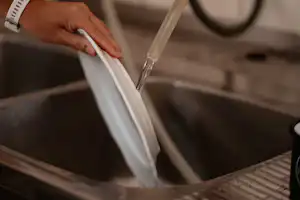 Hand gießt Wasser über Geschirr im Spülbecken, daneben ein Becher und Küchenutensilien auf einer Camping-Arbeitsfläche.