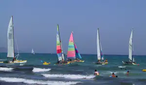 Segelboote auf dem Meer vor dem Strand, mehrere Jollen mit Segeln, Kinder im flachen Wasser und Wellen am Ufer.