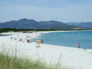 Sandstrand mit Liegen und Sonnenschirmen, im Hintergrund Wasser und Berge, am Ufer mehrere Personen und Boote.