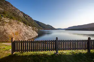 Blick vom Campingplatz auf einen See mit Holzzaun, Bergen und Uferstraße im Hintergrund.