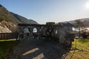 Grillplatz mit Steinterrasse und Steinmauer, Holzzaun und Feuerstelle; im Hintergrund Berge und grünes Gelände.