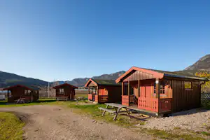 Mehrere Camping-Hütten aus Holz mit Veranda und Sitzbank, Kiesweg und Berge im Hintergrund.