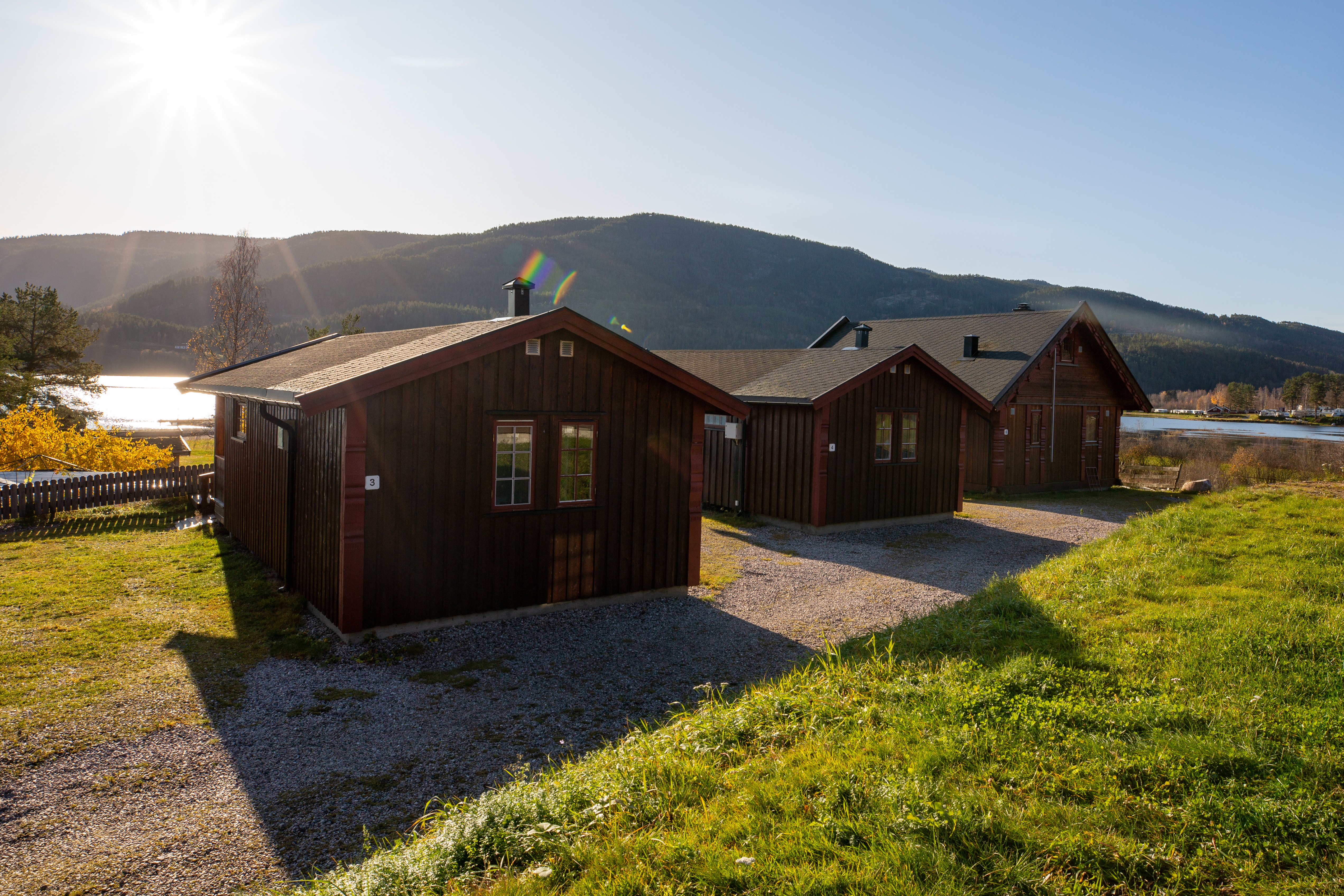 Holzhäuser auf einem Campingplatz mit gepflastertem Weg, Rasen und Blick auf bewaldete Berge in der Ferne.