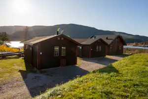 Holzhäuser auf einem Campingplatz mit gepflastertem Weg, Rasen und Blick auf bewaldete Berge in der Ferne.