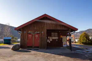 Holzgebäude am Campingplatz mit Eingangstür, Infotafel und überdachtem Bereich, daneben Mülltonnen und Weg.