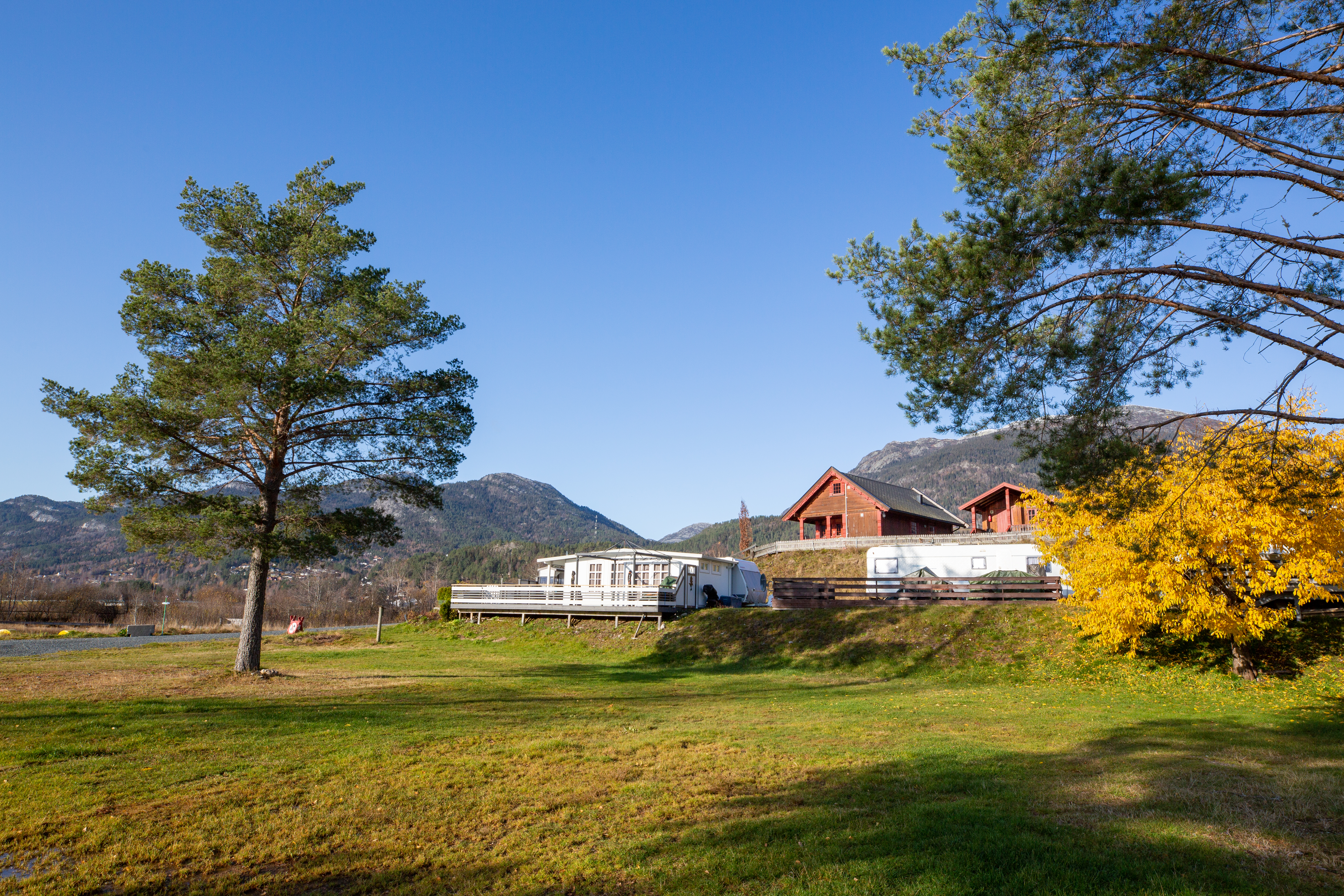 Campinghaus mit Holzfassade auf grünem Rasen, Zaun und Bäume im Vordergrund, Berge im Hintergrund.