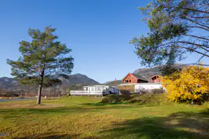 Campinghaus mit Holzfassade auf grünem Rasen, Zaun und Bäume im Vordergrund, Berge im Hintergrund.