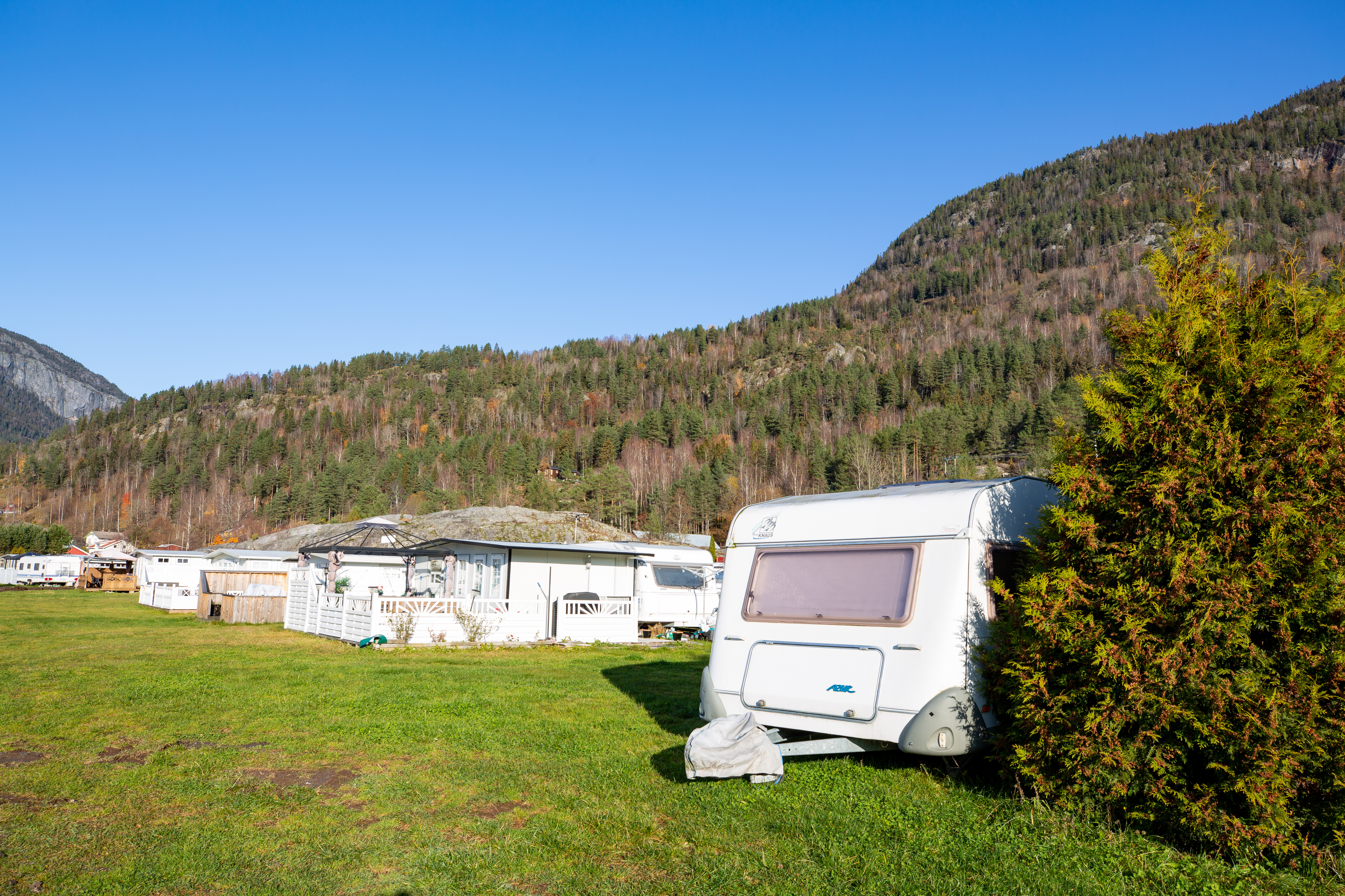 Wohnwagen auf einem grünen Campingplatz, daneben Hecken und weitere Wohnwagen, im Hintergrund Berge und ein Tal.
