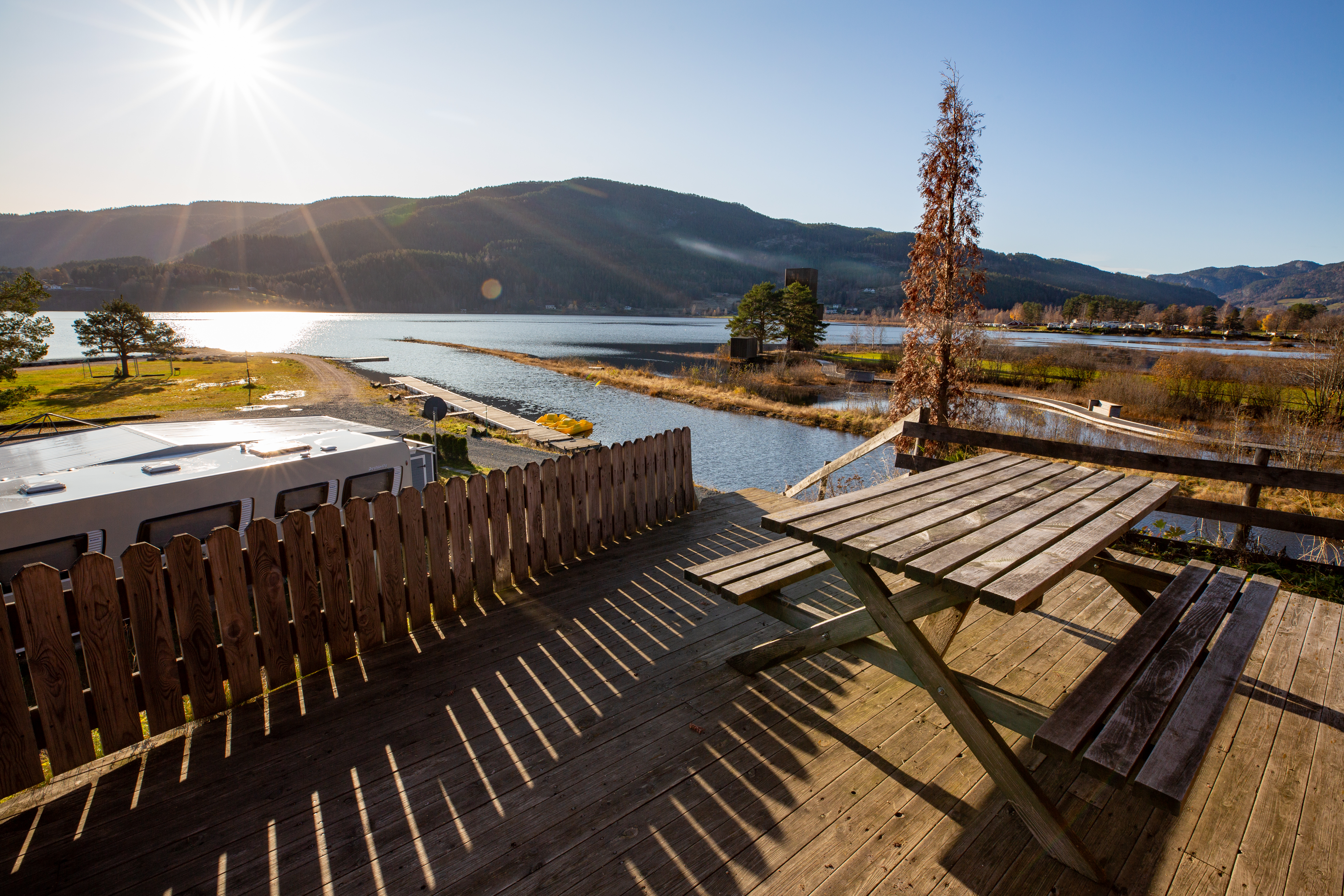 Holzterrasse mit Zäunen und Blick auf den See, im Hintergrund Berge und ein Campingplatz mit Wohnwagen-Stellplätzen.
