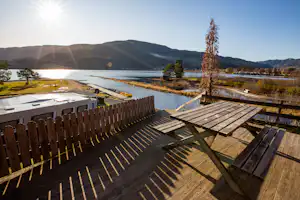 Holzterrasse mit Zäunen und Blick auf den See, im Hintergrund Berge und ein Campingplatz mit Wohnwagen-Stellplätzen.