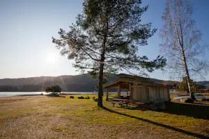 Holzhütte am Campingplatz am See, mit Sitzbank und Tisch, Grasfläche und Bäumen im Vordergrund.