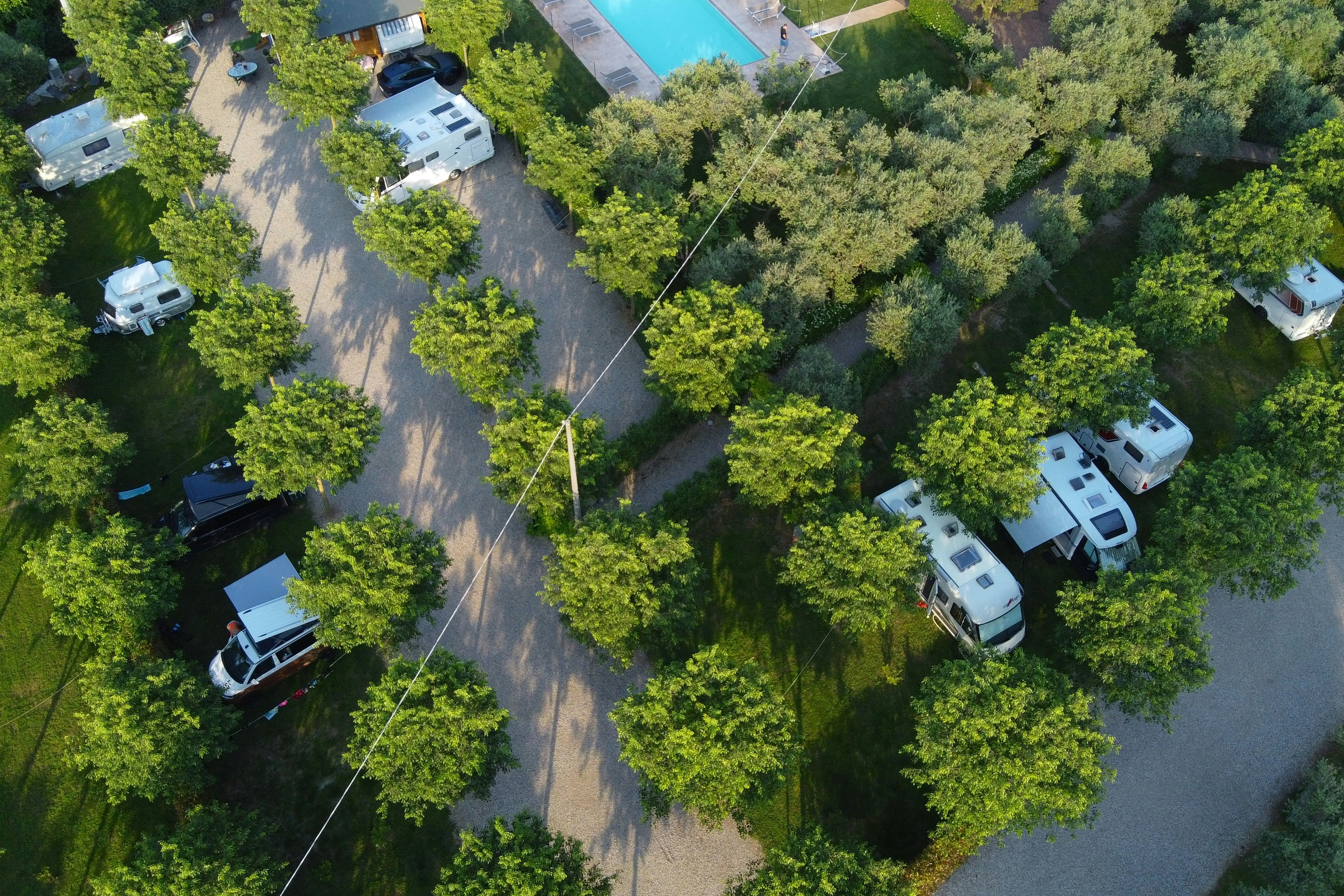 Luftaufnahme eines Campingplatzes mit Wohnmobilen und Wohnwagen auf Stellplätzen, umgeben von Bäumen und breiten Wegen.
