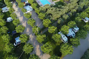 Luftaufnahme eines Campingplatzes mit Wohnmobilen und Wohnwagen auf Stellplätzen, umgeben von Bäumen und breiten Wegen.