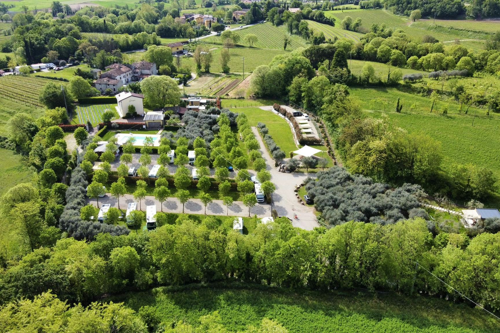 Luftaufnahme eines Campingplatzes mit Stellplätzen, Wohnwagen und Bäumen in grüner Landschaft, nahe einem kleinen Gebäude.
