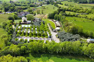 Luftaufnahme eines Campingplatzes mit Stellplätzen, Wohnwagen und Bäumen in grüner Landschaft, nahe einem kleinen Gebäude.