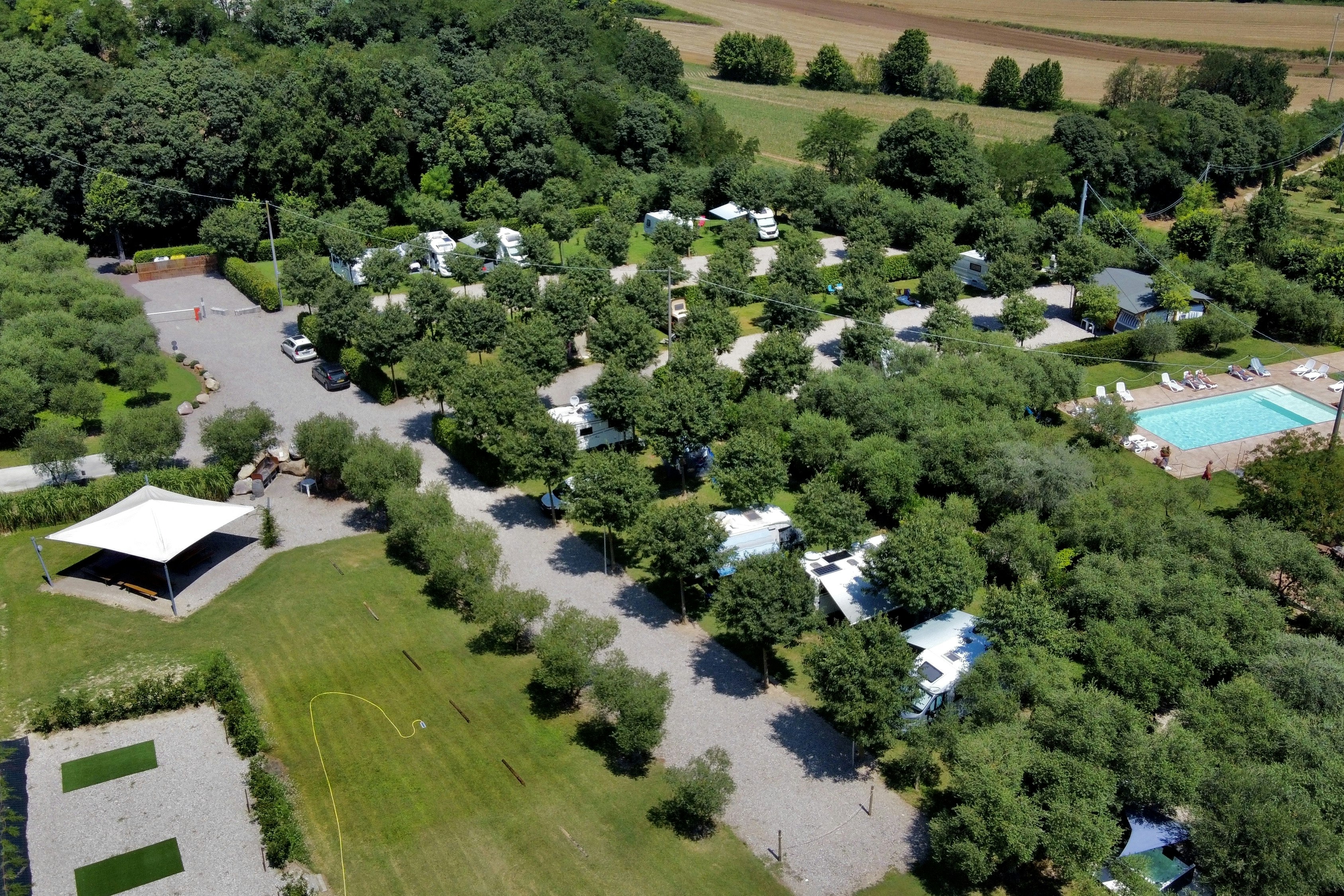Luftaufnahme eines Campingplatzes im Grünen mit Stellplätzen, Wegen, einem weißen Zelt und einem Pool.