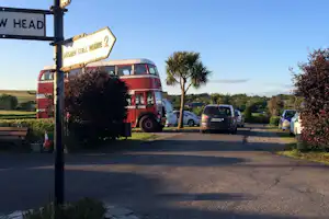 Rote Wohnmobile auf dem Campingplatz-Parkplatz, daneben Autos und ein Schild, im Hintergrund Felder und Bäume.